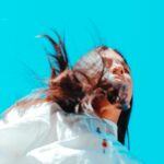Low angle view of a woman with windblown hair against a bright blue sky, showcasing fashion and freedom outdoors.