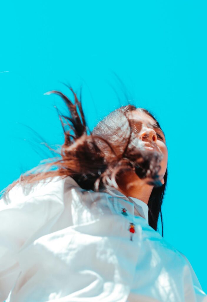 Low angle view of a woman with windblown hair against a bright blue sky, showcasing fashion and freedom outdoors.