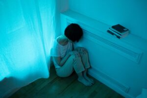 A woman in pajamas sits alone in a dimly lit room, expressing emotions of solitude.