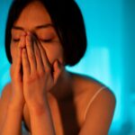 A woman with an emotional expression, covering her face in a serene bedroom setting.