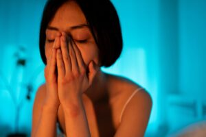 A woman with an emotional expression, covering her face in a serene bedroom setting.