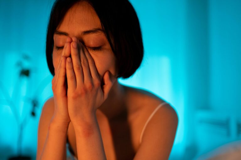 A woman with an emotional expression, covering her face in a serene bedroom setting.