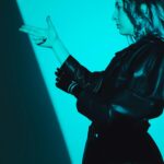 Side view of a woman in a leather coat making a hand gesture, captured in blue lighting.