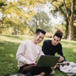 Full length content ethnic male students in casual outfits browsing laptop while sitting on meadow with copybooks and preparing for exam in green park