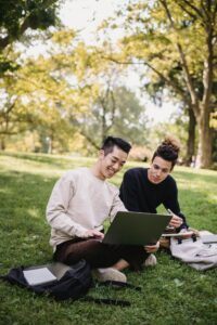 Full length content ethnic male students in casual outfits browsing laptop while sitting on meadow with copybooks and preparing for exam in green park