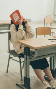 A student playfully hiding their face with a book in a classroom, sitting at a desk.