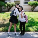 Two friends hugging outside a school building on a sunny day, both wearing backpacks.