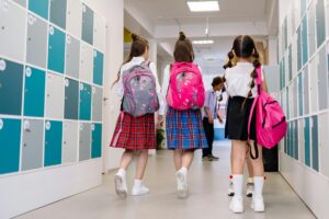 Three school girls with backpacks walking in a school corridor lined with lockers.