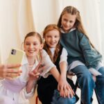 Three teenage girls smiling while taking a selfie together indoors.