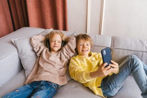 Two children laughing and posing for a selfie on a cozy couch, capturing a joyful moment of friendship and fun.
