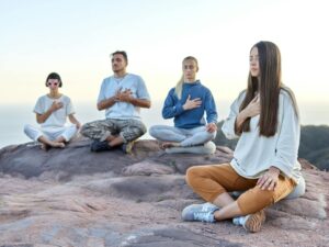 A diverse group of adults meditating outdoors on a rocky landscape with a serene background.