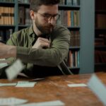 A bearded man throws papers in a library while looking frustrated, sitting at a wooden table.