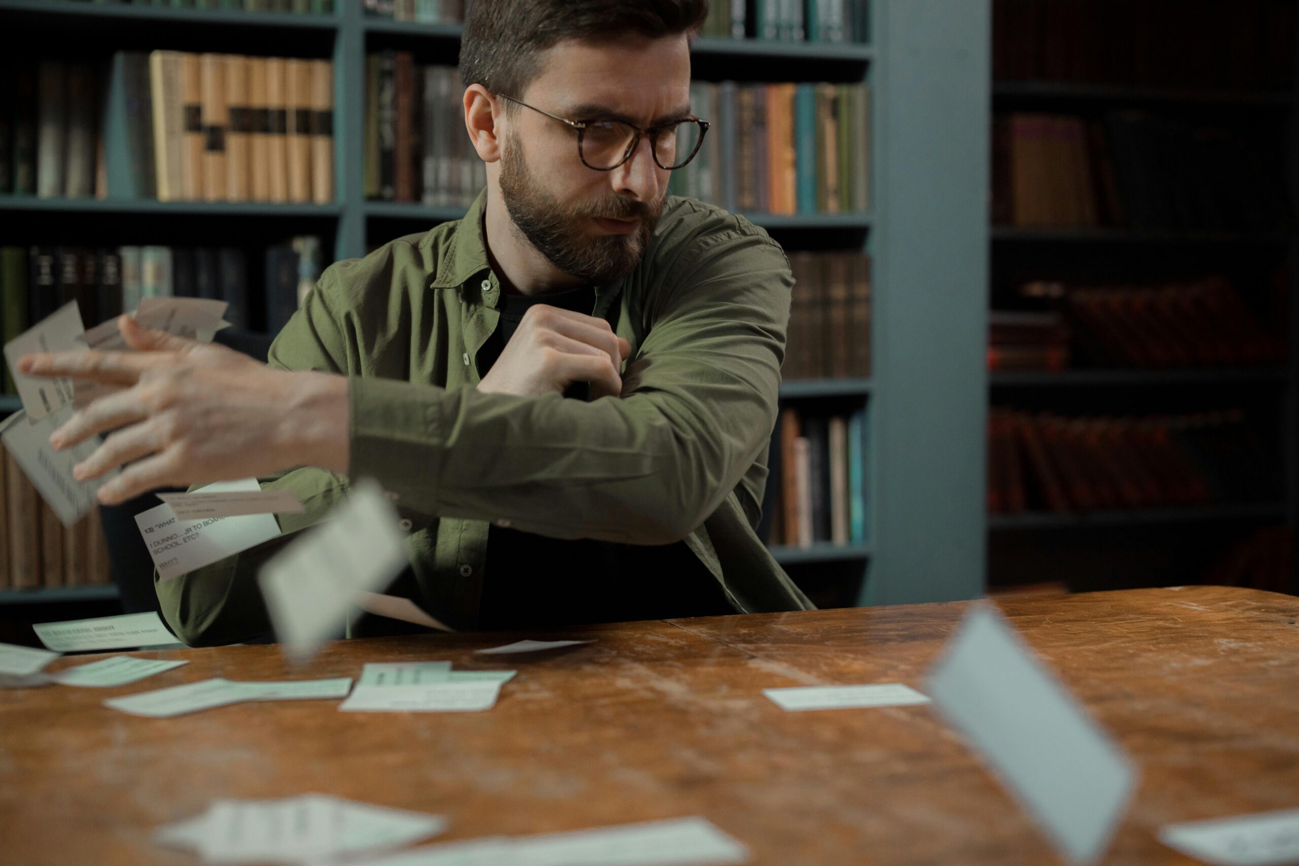 A bearded man throws papers in a library while looking frustrated, sitting at a wooden table.