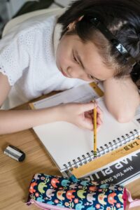 Young Asian girl diligently studying indoors, focused on her homework with stationery.