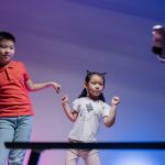 Two children joyfully dance with a robot in a colorful indoor setting.