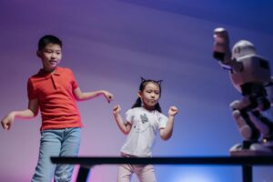 Two children joyfully dance with a robot in a colorful indoor setting.