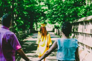 A graduate in a yellow gown walks ahead as parents hold hands outdoors, celebrating a new chapter.