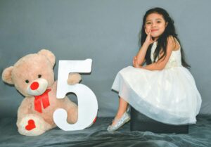 Charming portrait of a girl in a white dress celebrating her fifth birthday with a teddy bear.