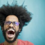 Dynamic portrait capturing a man with afro hair and eyeglasses shouting energetically in studio setting.