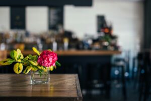 A vibrant pink flower in a glass vase on a wooden table in a modern café with a blurred background.