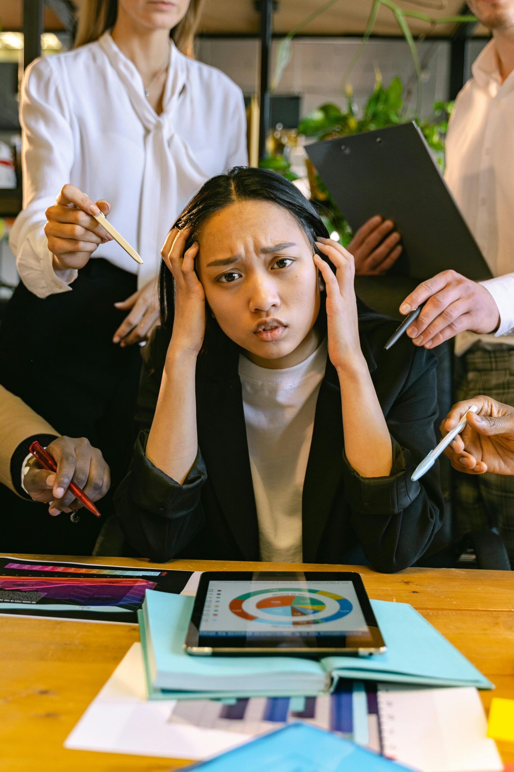 Stressed Asian businesswoman surrounded by demanding colleagues in a busy office setting.