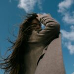 Low-angle shot of a woman with long hair in a brown coat looking over her shoulder against a bright sky.