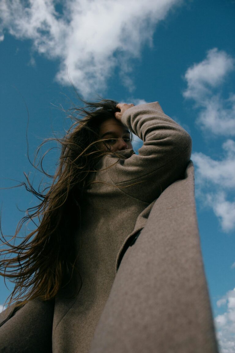Low-angle shot of a woman with long hair in a brown coat looking over her shoulder against a bright sky.