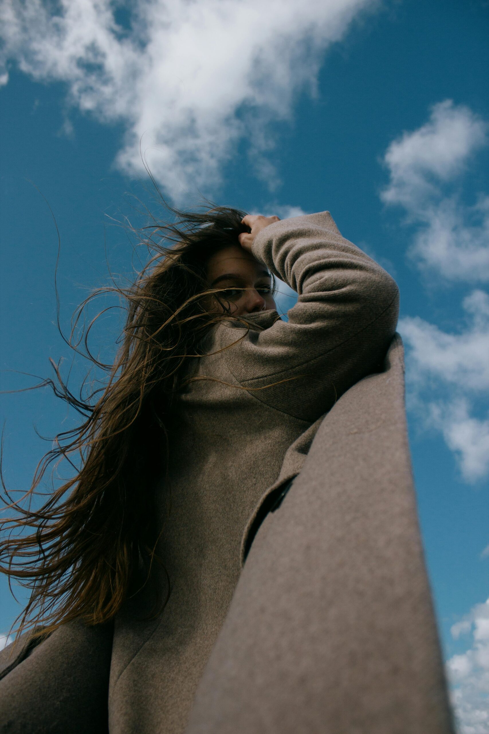 Low-angle shot of a woman with long hair in a brown coat looking over her shoulder against a bright sky.