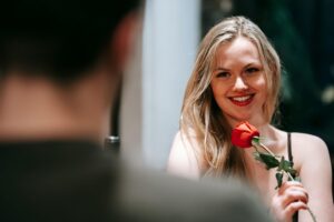 Young couple in elegant clothes sitting in light restaurant while enjoying date with red rose and looking at each other