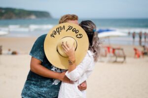 A couple shares a romantic kiss on a sunny beach, hidden behind a wide-brimmed hat.