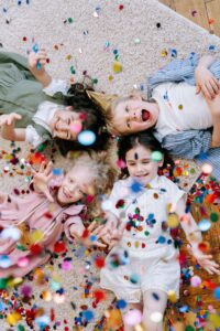Four happy children enjoy a party, lying on the floor with colorful confetti showering around them.