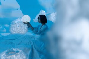 雲が立ち込める中で、光り輝く球体を手に持った女性の幻想的なイメージ。夢のような雰囲気を醸し出している。