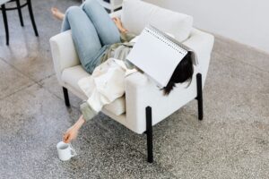 肘掛け椅子に横たわり、顔に本を乗せ、コーヒーを持っている女性。カジュアルでリラックスした雰囲気。