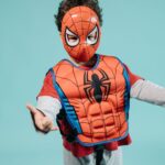 Child wearing a Spiderman costume and mask striking a playful superhero pose in a studio setting.