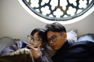 A father and son wearing glasses enjoy a relaxing moment together, using a smartphone indoors.