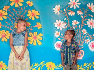 Two children smiling in front of a vivid mural of colorful flowers. Bright and joyful moments.