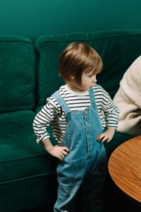 Adorable child in denim overalls with hands on waist, standing by a green couch indoors.